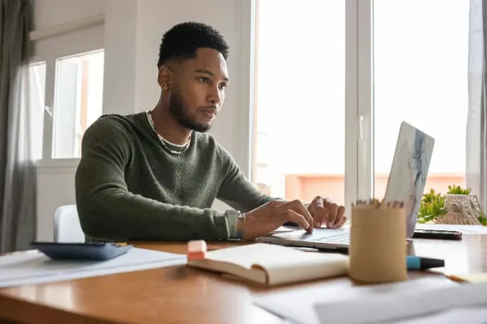 A focused young man working on a laptop at a wooden desk.