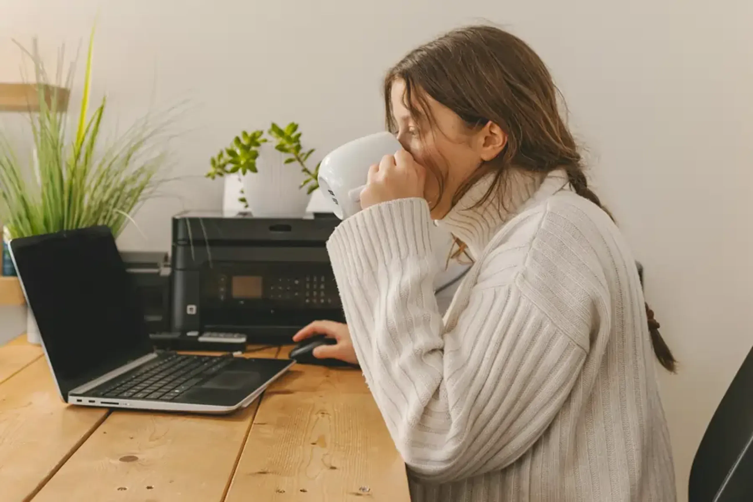 Woman drinking coffee while working on a laptop at a desk.