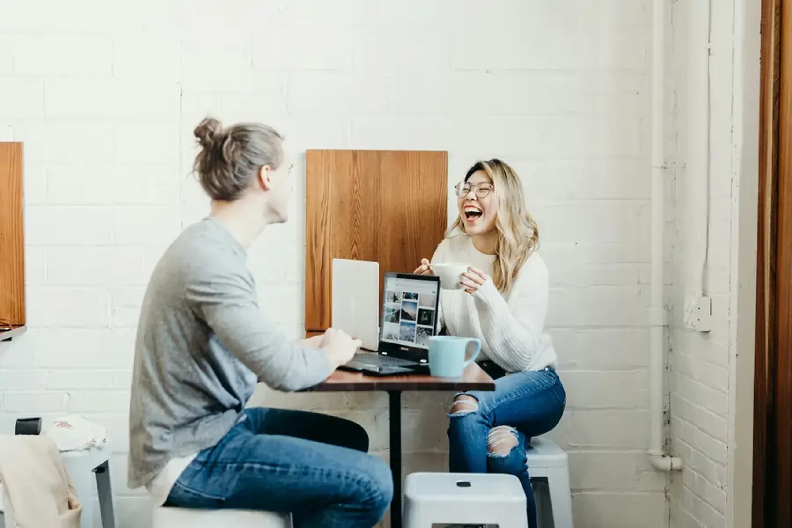 Two people laughing and talking while working on laptops at a table.