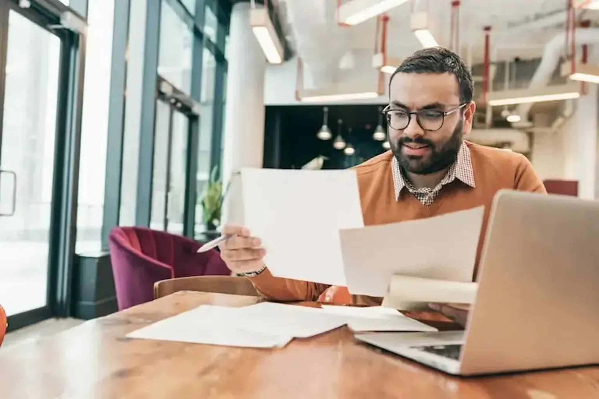 Man reviewing documents while working on a laptop at a desk.