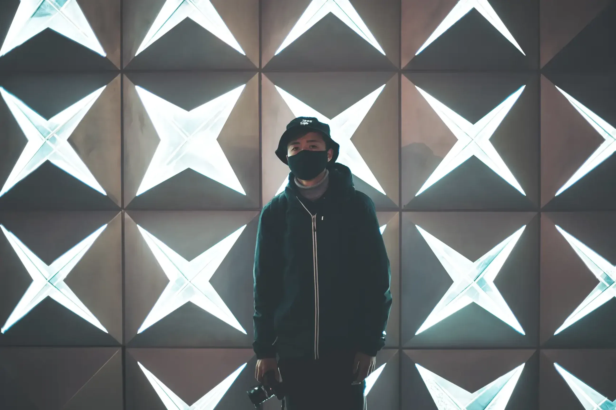 Person wearing a mask standing in front of a geometric wall of illuminated X-shaped lights.